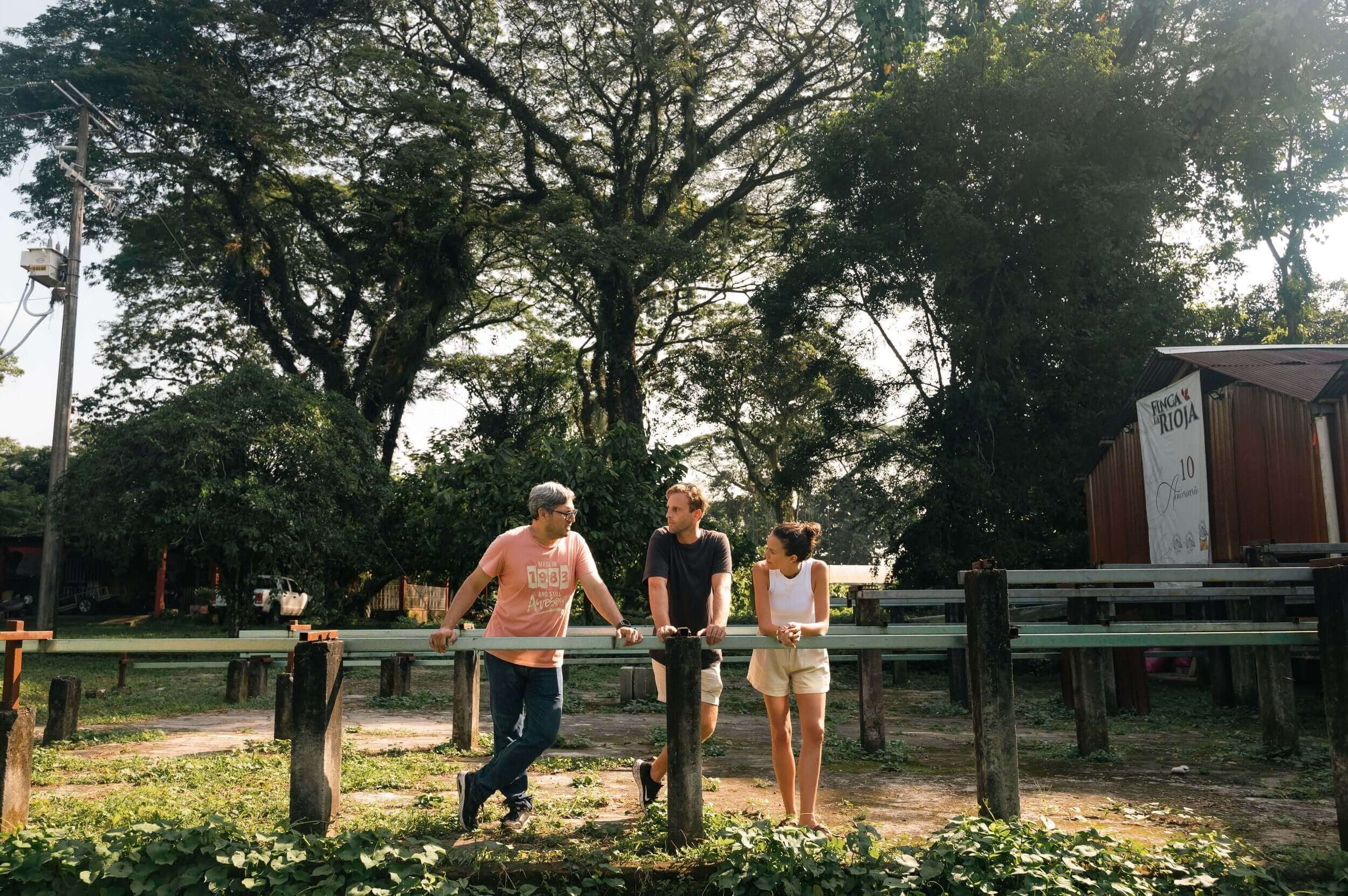 Three people conversing outdoors in a lush Mexican village, surrounded by greenery and a rustic setting.