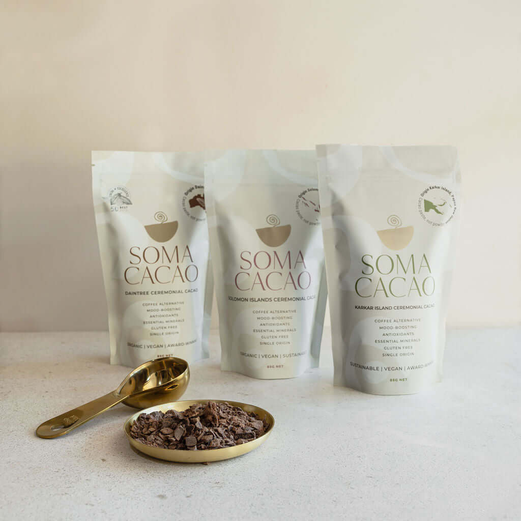 Three Soma Cacao bags with ceremonial cacao next to a golden spoon and dish on a light background.