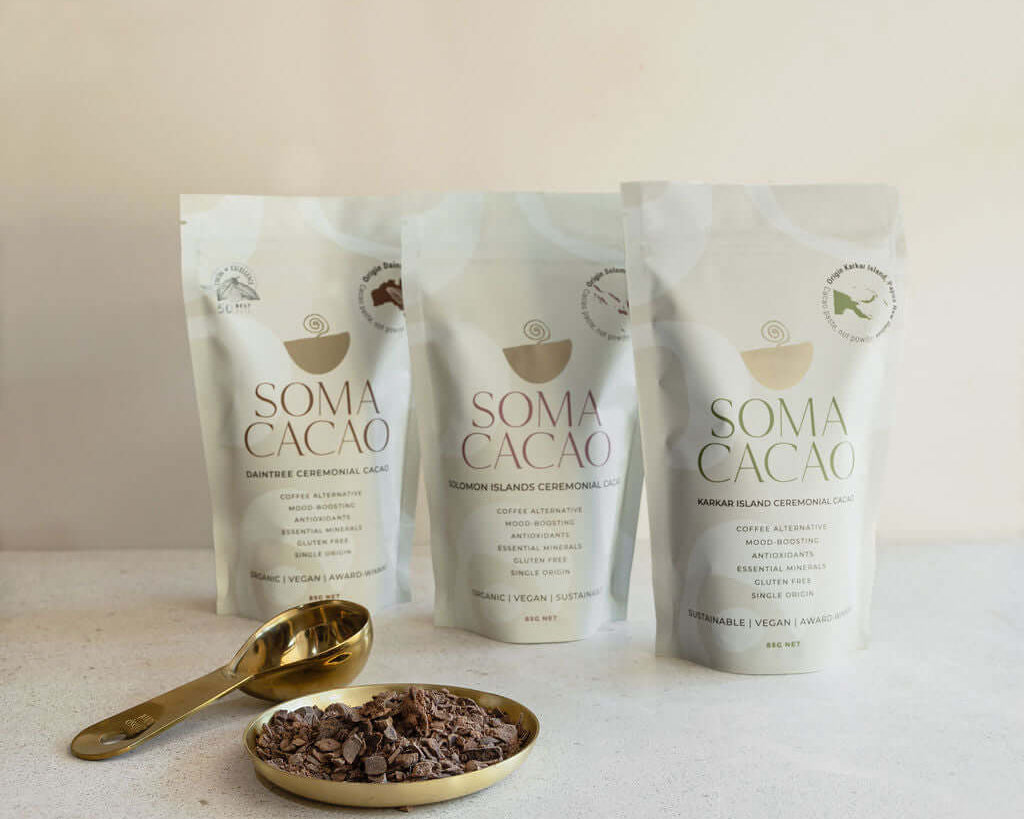 Three Soma Cacao bags with ceremonial cacao next to a golden spoon and dish on a light background.