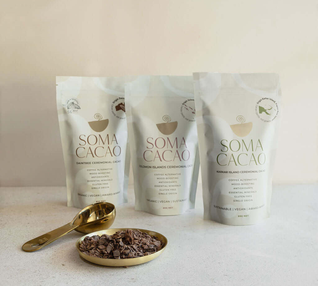 Three Soma Cacao bags with ceremonial cacao next to a golden spoon and dish on a light background.