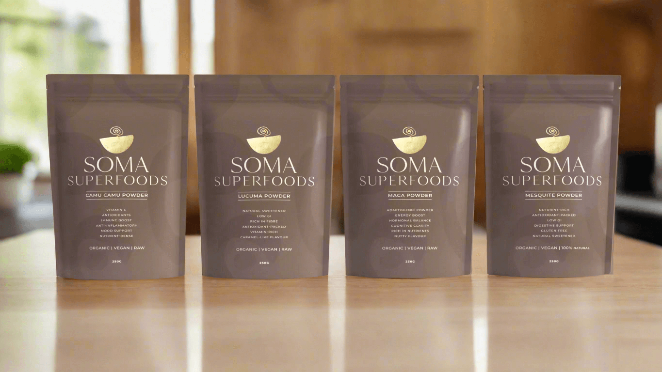 Four packs of SOMA Superfoods: Camu Camu, Lucuma, Maca, and Mesquite powders on a wooden table.