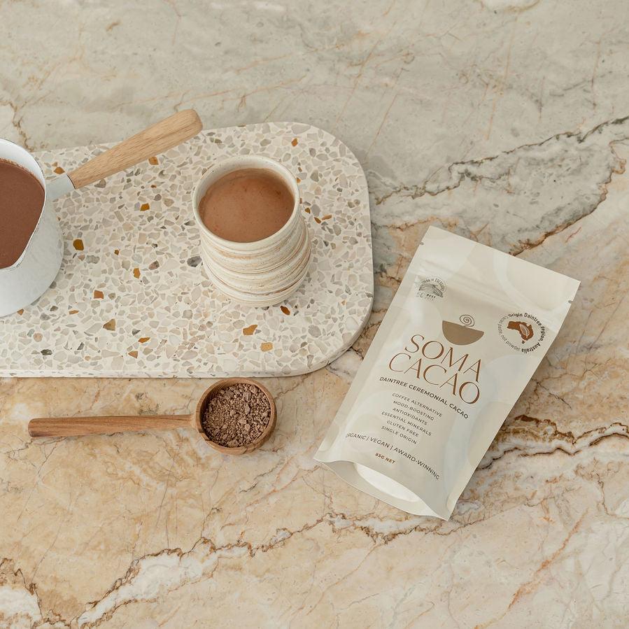 Daintree ceremonial cacao packet on a marble surface with a cup of cacao drink and a wooden spoon for serving.