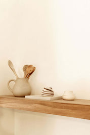 Minimalist wooden shelf with ceramic mugs and wooden spoons in a jug, styled on a neutral background.