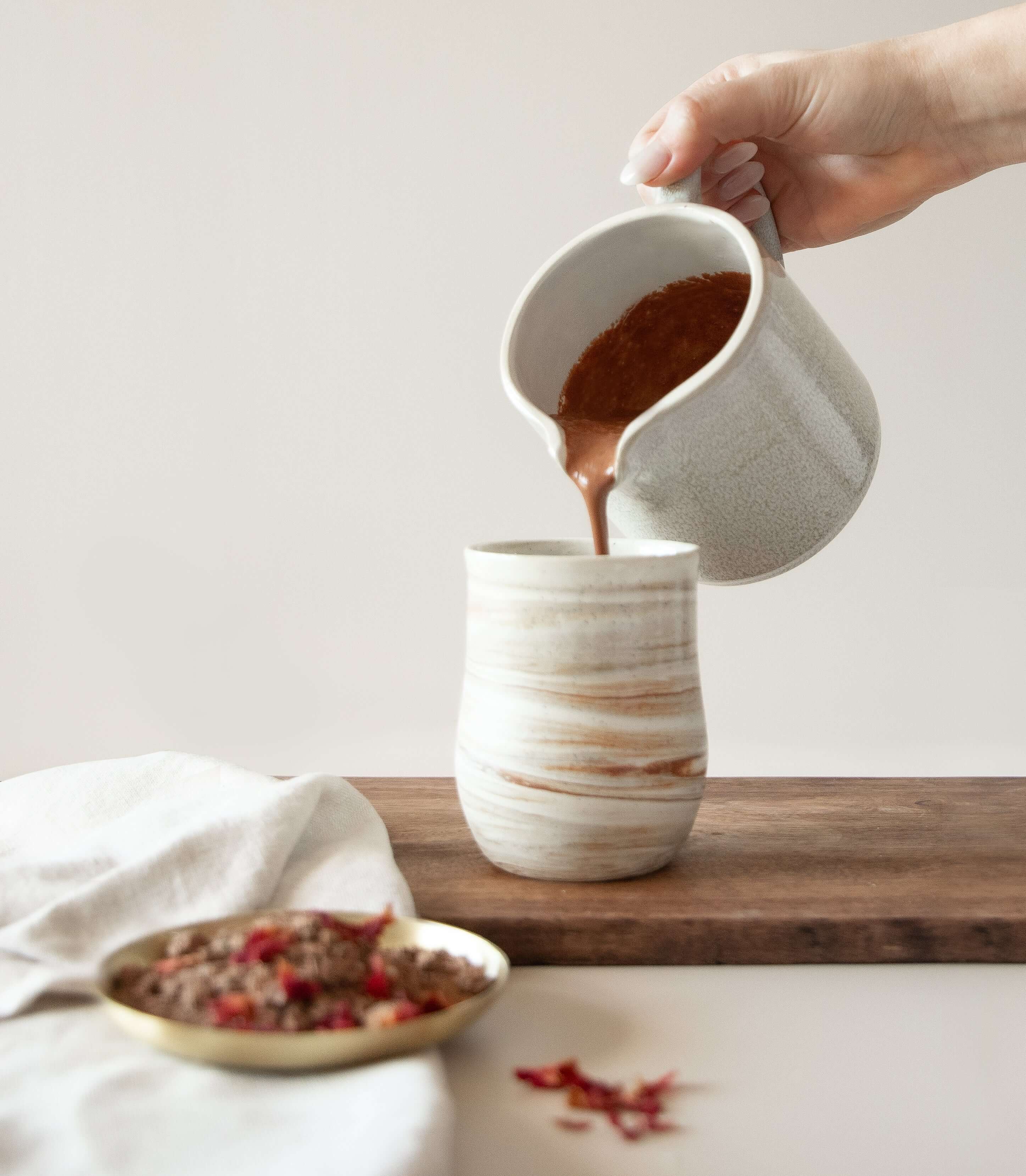 Hand pouring rich ceremonial cacao into a textured cup, showcasing premium quality and warmth in a serene setting.