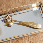 Brass tea scoop with loose tea leaves on mirrored tray, enhancing ceremonial cacao ritual with style and function.